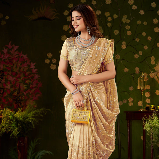 A woman models a beige and gold embroidered saree with a matching embellished blouse, standing in a decorated indoor setting with a dark green floral wall and patterned rug.