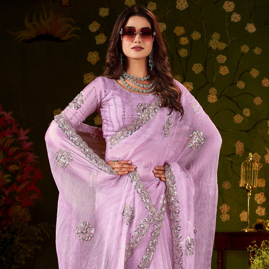 A woman models a lavender purple saree with intricate silver embroidery, featuring a matching embroidered blouse and a sheer dupatta, set against a dark, ornate background with floral patterns.