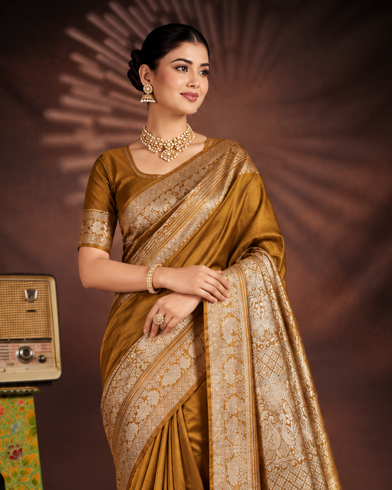 A woman models a bronze-gold saree with intricate beige and gold zari embroidery, standing in front of a dark brown studio backdrop. She is accessorized with a gold necklace and earrings, and a small vintage radio and decorative stool are placed to her left.