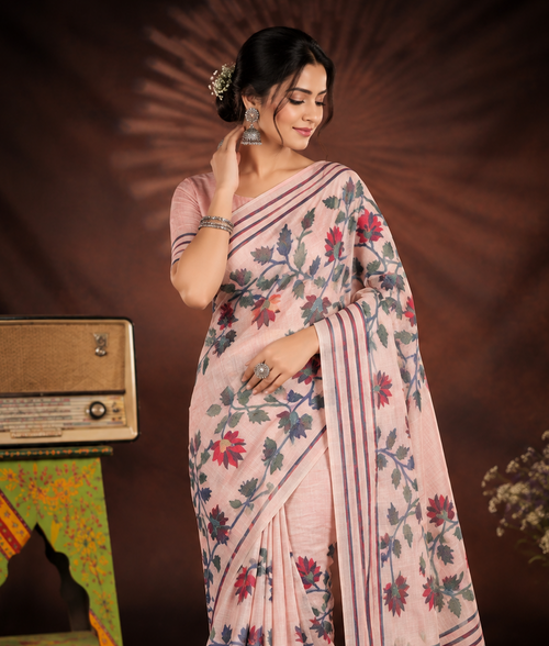 A woman models a light pink saree with a floral print in red, blue, and green, styled with a matching pink blouse. The scene is set against a dark brown studio backdrop with vintage props, including a small radio on a green stand and a vase of flowers.