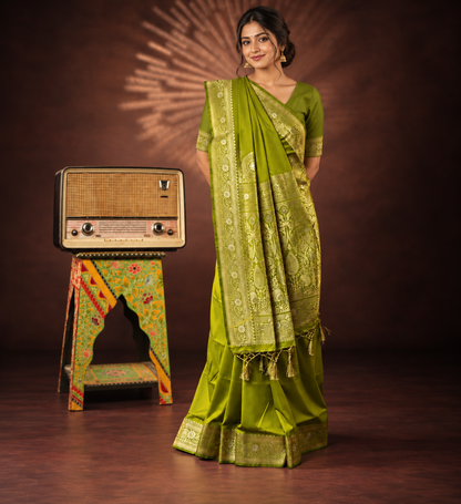 A woman models an olive green saree with intricate gold zari embroidery, standing next to a vintage radio on a decorated stand against a dark brown studio backdrop.