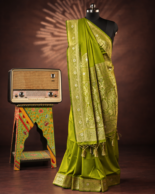 A green silk saree with intricate gold zari embroidery and border patterns, draped on a black mannequin. The mannequin is set against a dark brown studio background, with a vintage radio on a decorated stand to the left.