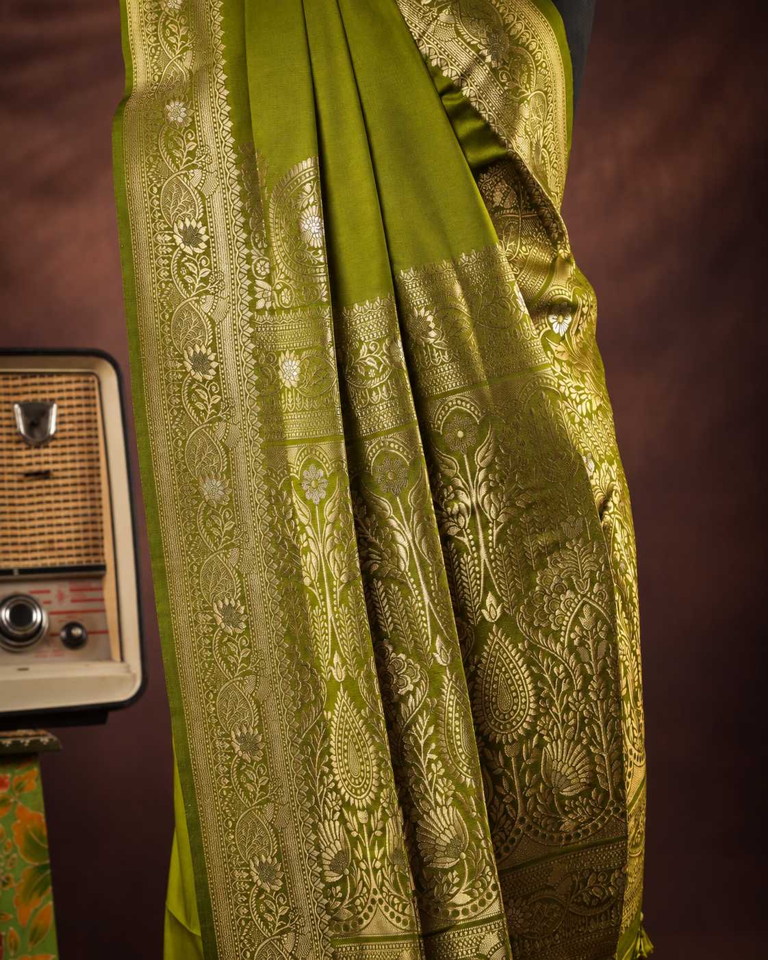 an olive green silk saree with intricate gold zari woven patterns, draped elegantly next to a vintage radio against a dark brown background.