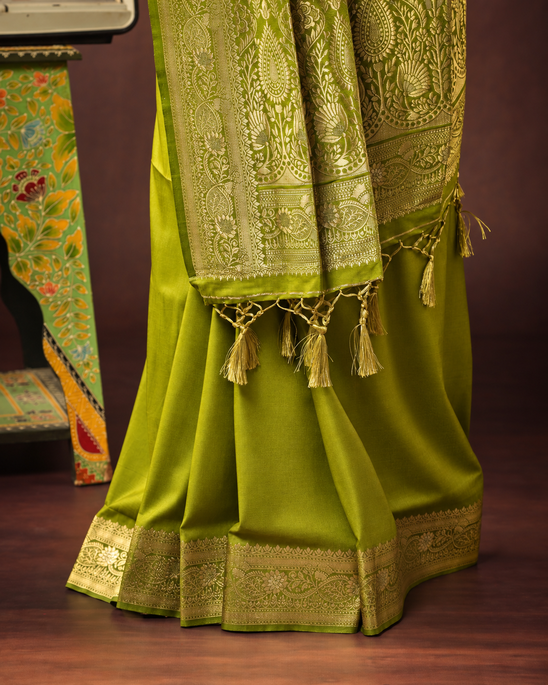 An olive green saree with intricate gold zari embroidery, featuring a wide border, tassels, and a matching dupatta draped over the skirt, set against a dark brown background.