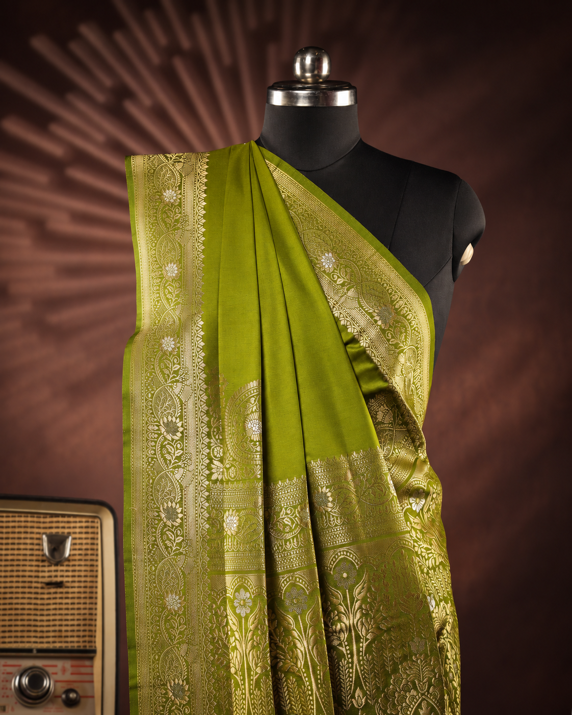 An olive green silk saree with intricate gold zari embroidery, draped on a black mannequin torso. The background is a dark brown studio backdrop with a sunburst pattern, and a vintage radio is partially visible on the bottom left.
