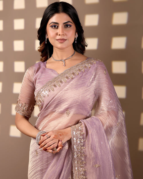 A woman wearing a lavender purple saree with silver embellished borders and a matching beads work blouse, standing against a brown background with a geometric pattern.